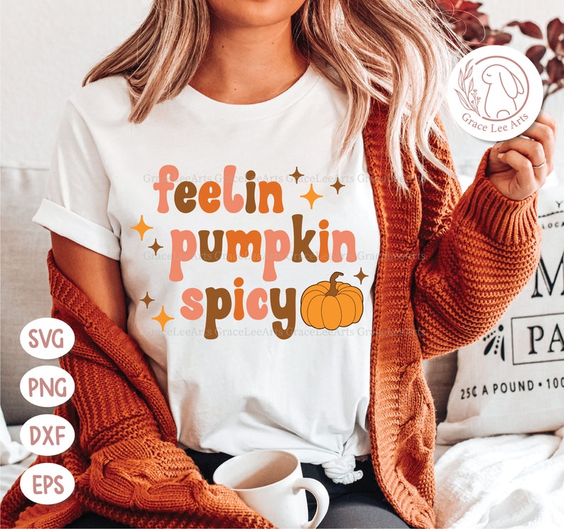 May include: White t-shirt with the text "feelin' pumpkin spicy" in brown, pink, and orange letters. A small orange pumpkin with a green stem is next to the text.