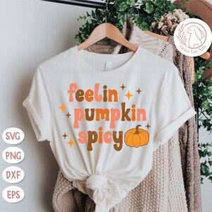 May include: A white t-shirt with the text "feelin' pumpkin spicy" in a retro font. The text is in shades of orange, brown, and pink. A small orange pumpkin is to the right of the text.