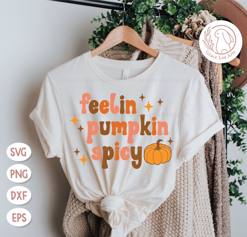 May include: A white t-shirt with the text "feelin' pumpkin spicy" in a retro font. The text is in shades of orange, brown, and pink. A small orange pumpkin is to the right of the text.