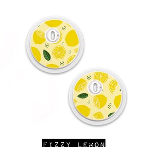 May include: Two round medical devices with a lemon pattern on a yellow background. The design includes whole and sliced lemons, green leaves, and small black dots. The text "FIZZY LEMON" is displayed in a black rectangle.
