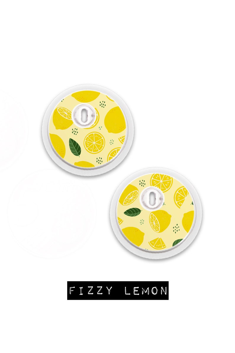 May include: Two round medical devices with a lemon pattern on a yellow background. The design includes whole and sliced lemons, green leaves, and small black dots. The text "FIZZY LEMON" is displayed in a black rectangle.