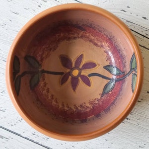 May include: A round, terracotta-colored bowl with a floral design. The interior features a central purple flower with yellow accents, surrounded by a reddish-brown and tan background. Green leaves and stems extend from the flower.