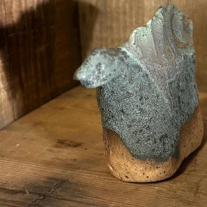 May include: A small ceramic sculpture of a creature with a textured, mottled blue-green glaze on the upper portion and a smooth, speckled tan base. The object features a prominent, jagged dorsal ridge and a rounded head, crafted from clay.