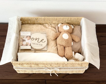 Può includere: Un cesto intrecciato pieno di articoli per bambini. Include un orsacchiotto, una coperta, un disco di legno con la scritta "one month" e altri elementi essenziali per bambini. Il cesto è foderato con carta bianca.