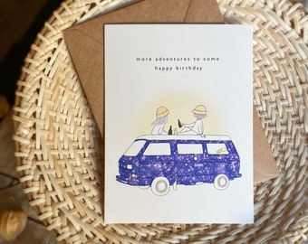 May include: A birthday card with a blue and white illustration of two people in a van. The text on the card reads "more adventures to come happy birthday".