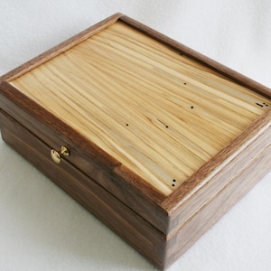 May include: A rectangular wooden box with a light-colored, striped top and dark brown sides. The box has a brass-colored clasp and is closed. The wood grain is visible on the top and sides, showcasing the natural beauty of the material.