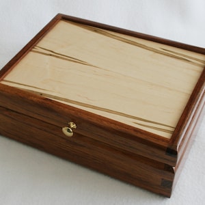 May include: A rectangular wooden box with a light-colored top and dark brown sides. The top has a light wood center with dark wood trim and gold accents. A small, round, gold-colored knob is on the front. The box is closed.