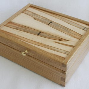 May include: A rectangular wooden box with a light brown finish. The lid features a sunburst design with alternating light and dark wood strips. A small, gold-colored clasp secures the box.