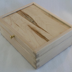 May include: A rectangular wooden box with a light-colored finish and a contrasting grain pattern on the lid. The box features dovetail joints and a small, gold-colored latch. The box is made of wood.