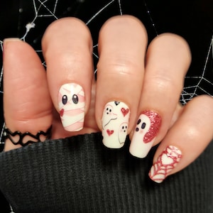 May include: Close-up of a hand with five white nails painted with Halloween-themed nail art. The nails feature ghosts, hearts, and a mummy design. The nail art is done in white, pink, and red.