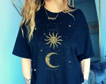 May include: A black t-shirt with a gold sun and moon graphic and the text "live by the sun love by the moon". The shirt has a relaxed fit and is decorated with small gold stars.