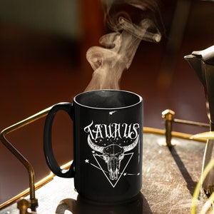 May include: Black ceramic mug with a white design of a bull skull and the word "TAURUS" in a stylized font. Steam rises from the mug.