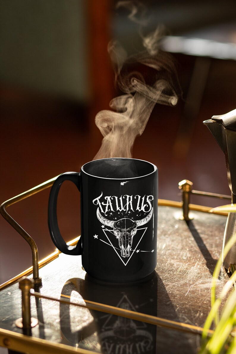 May include: Black ceramic mug with a white design of a bull skull and the word "TAURUS" in a stylized font. Steam rises from the mug.