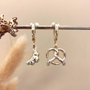 Puede incluir: Pendientes de aro dorados con colgantes. Un pendiente presenta un dije de croissant, y el otro tiene un dije de pretzel. Los pendientes se exhiben sobre un fondo suave y neutro, resaltando los detalles de la joyería.
