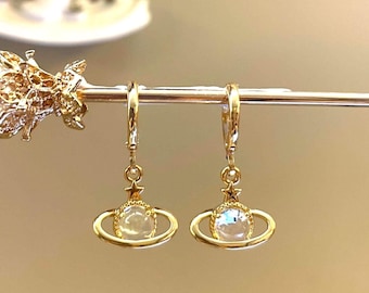 Puede incluir: Un par de pendientes de aro dorados con un dije colgante que presenta un orbe de cristal transparente rodeado por un anillo dorado y una pequeña estrella dorada.