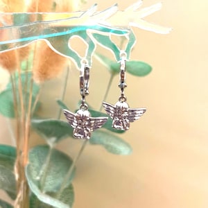 May include: A pair of silver hoop earrings with angel charms. The angels are facing forward with their wings spread out.