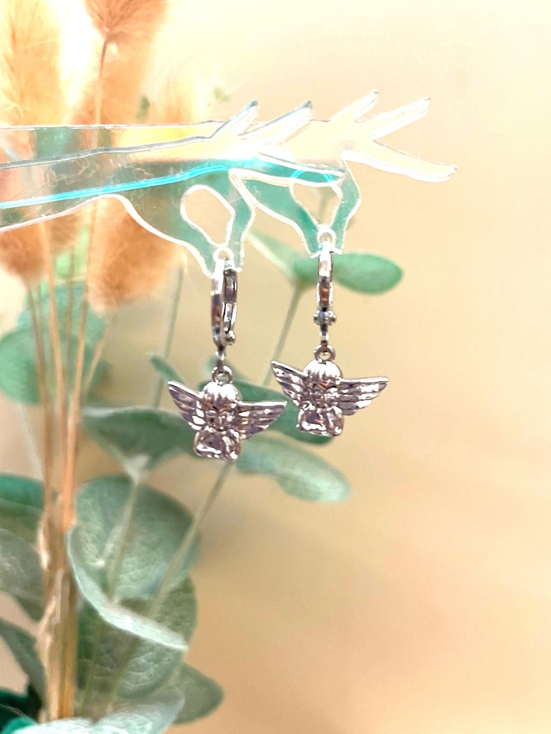 May include: A pair of silver hoop earrings with angel charms. The angels are facing forward with their wings spread out.