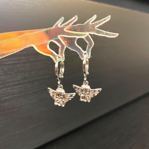May include: A pair of silver hoop earrings with angel charms. The angels are sitting with their wings spread out.