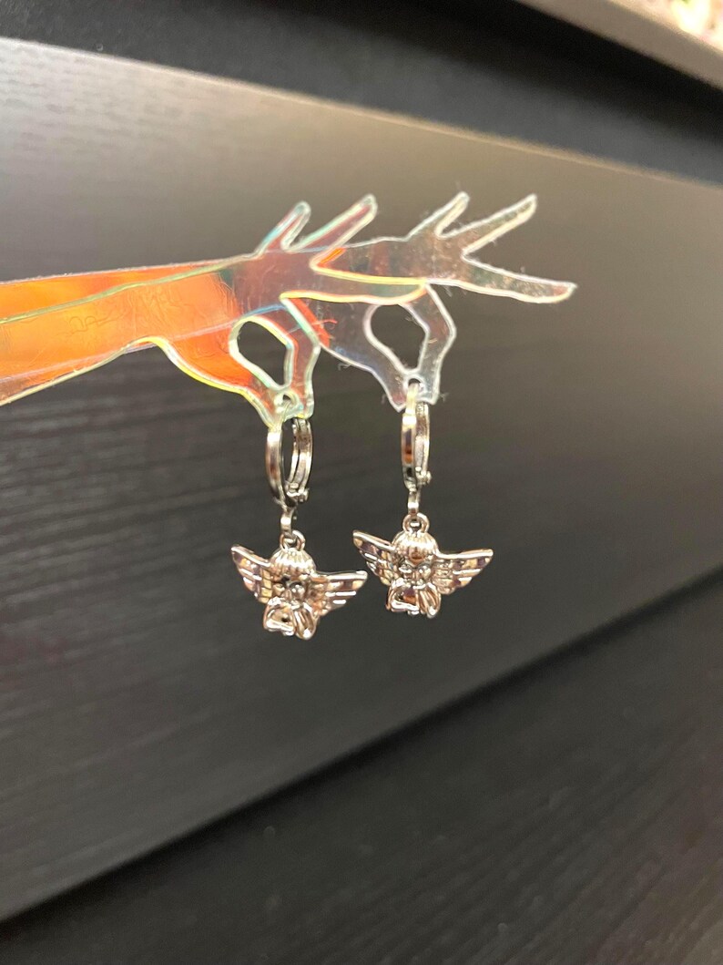 May include: A pair of silver hoop earrings with angel charms. The angels are sitting with their wings spread out.