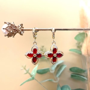Op de afbeelding: Een paar goudkleurige oorringen met een vierbladige bloemvorm. Elk bloemblad is een dieprode edelsteen, omringd door kleine, heldere kristallen. De oorbellen worden gepresenteerd op een goudkleurige staaf.