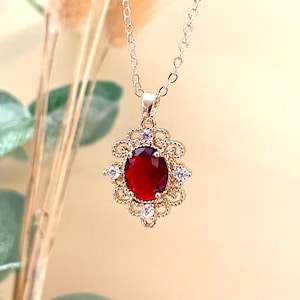 Puede incluir: Un collar con colgante dorado con una gran gema roja ovalada. La gema está engastada en un engaste de filigrana dorada decorativa con pequeñas piedras de acento transparentes. La cadena del collar también es dorada.