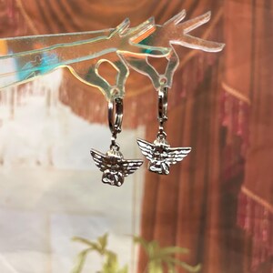 May include: A pair of silver hoop earrings with angel charms. The angels are depicted with wings and a halo.