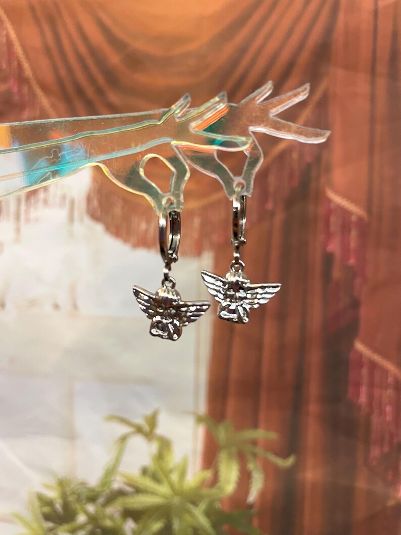 May include: A pair of silver hoop earrings with angel charms. The angels are depicted with wings and a halo.