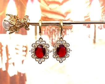 May include: A pair of gold earrings with red oval-shaped gemstones surrounded by smaller clear gemstones. The earrings have a delicate, ornate design.