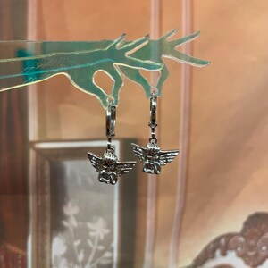 May include: A pair of silver hoop earrings with a small angel charm hanging from each hoop.