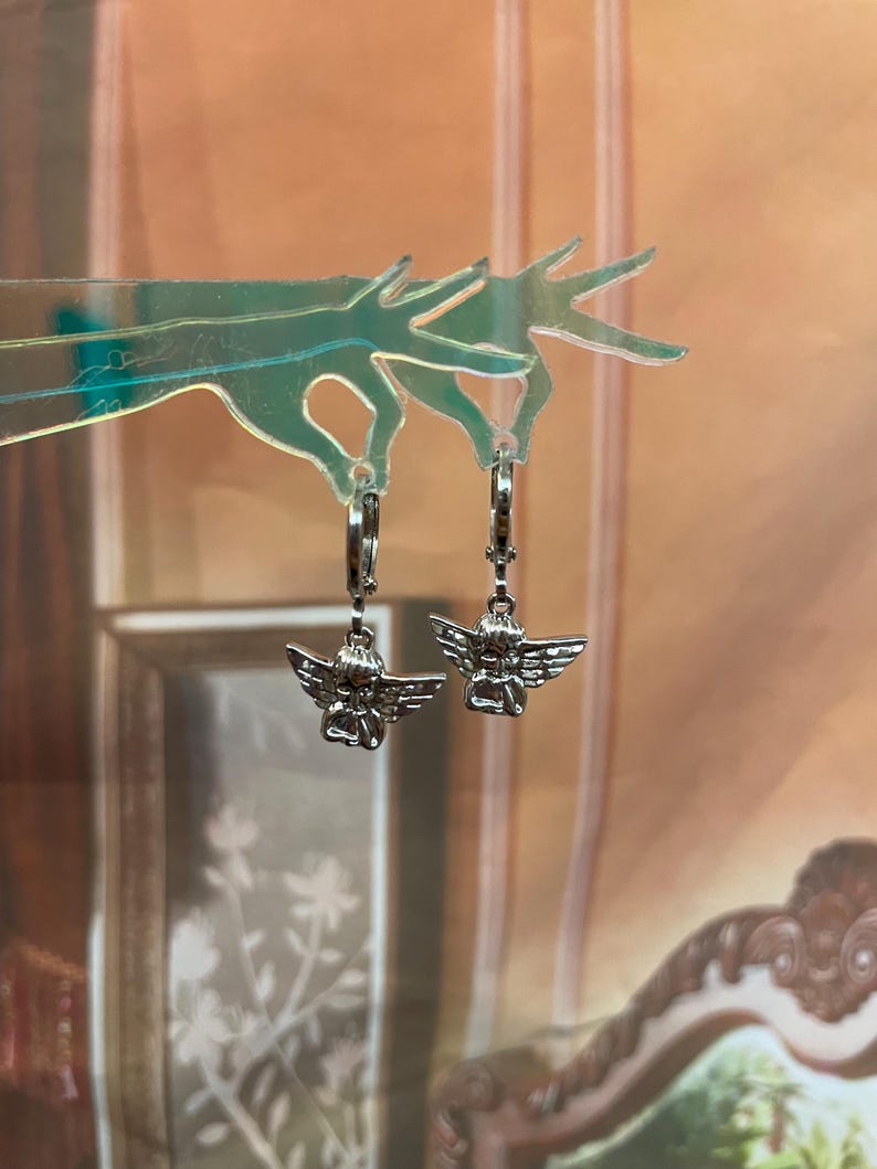 May include: A pair of silver hoop earrings with a small angel charm hanging from each hoop.