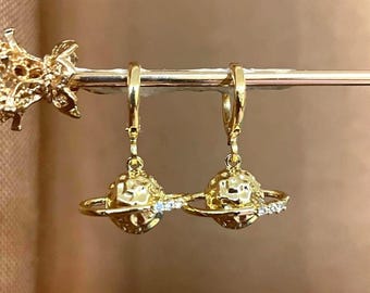 Puede incluir: Pendientes de aro dorados con una esfera texturizada y un anillo de pequeños diamantes alrededor de la esfera, que se asemeja a un planeta con anillos.
