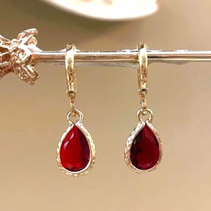 Op de afbeelding: Een paar gouden creolen met hangende rode druppelvormige stenen. De stenen zijn gezet in een gouden frame met een getextureerd ontwerp.