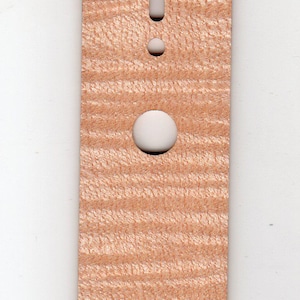 May include: A light brown wooden guitar part with a textured surface. It has a long, rectangular shape with rounded ends and several holes. There is a long, narrow slot in the centre. The part is likely a guitar neck plate.