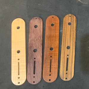 Może przedstawiać: Cztery osłony pickupa do gitary wykonane z różnych rodzajów drewna. Osłony pickupa są wszystkie tego samego rozmiaru i kształtu, ale mają różne usłojenia i kolory. Osłony pickupa są ułożone w rzędzie od lewej do prawej. Osłona pickupa po lewej stronie jest wykonana z jasnego drewna w jasnobrązowym kolorze. Osłona pickupa w środku jest wykonana z ciemnego drewna w brązowym kolorze. Osłona pickupa po prawej stronie jest wykonana z jasnego drewna w brązowym kolorze.