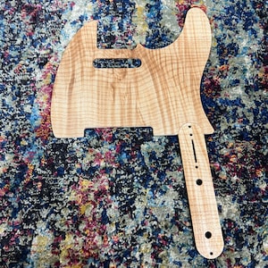 Peut inclure: Un corps de guitare en bois naturel de couleur brun clair avec un motif de grain de bois. Le corps est façonné comme une guitare électrique traditionnelle avec un pan coupé pour accéder aux frettes supérieures. Le corps a un manche qui lui est attaché, également en bois naturel.
