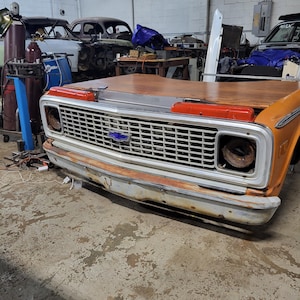 May include: A close-up of the front of an orange vintage pickup truck with a wooden bed. The truck has a chrome grille and a white bumper. The truck is in a garage with other vehicles in the background.