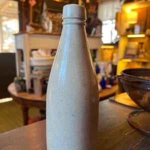 May include: A vintage, off-white stoneware bottle with a narrow neck and rounded body. The bottle has a slightly textured surface and a simple, classic design. It is likely an antique or a reproduction of an antique bottle.