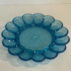 May include: A turquoise glass egg serving platter with a scalloped edge. The platter has 12 individual egg cup holders around the edge and a textured center. The glass has a slight iridescence.