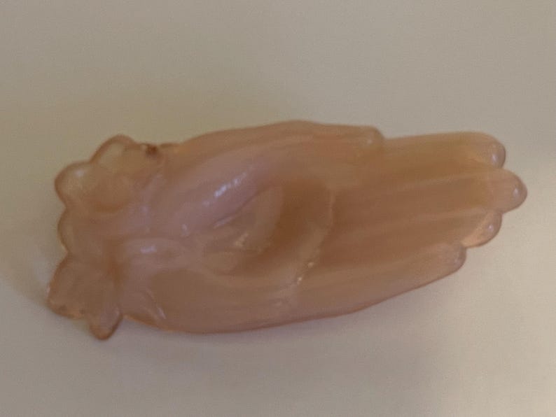 May include: A pale pink glass hand-shaped dish. The dish is shaped like a hand with the fingers curled inward.