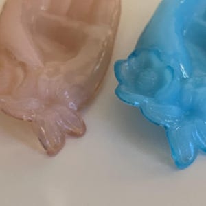 May include: Two glass hand-shaped dishes with floral details. One dish is light pink and the other is light blue.