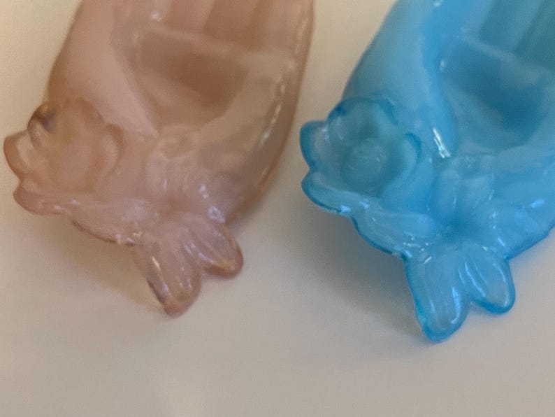 May include: Two glass hand-shaped dishes with floral details. One dish is light pink and the other is light blue.