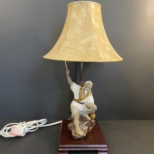 May include: A decorative table lamp featuring a figurine of an elderly man in traditional attire, holding a fishing rod. The lamp has a beige lampshade and sits on a dark wooden base. The lamp is connected to a white electrical cord.