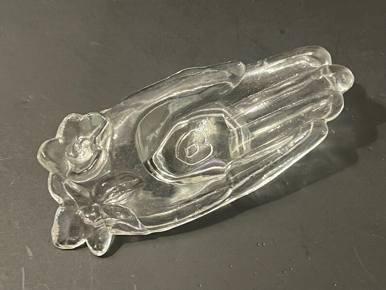 May include: A clear glass hand-shaped dish with a heart-shaped indentation in the palm. The dish has a floral design around the base.
