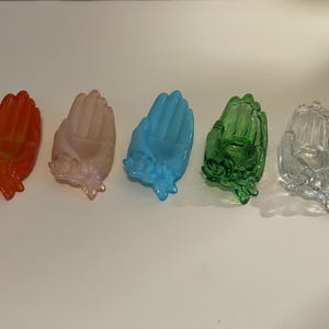 May include: Five glass hand-shaped trinket dishes in various colors. The dishes are arranged in a row from left to right: orange, pink, blue, green, and clear.