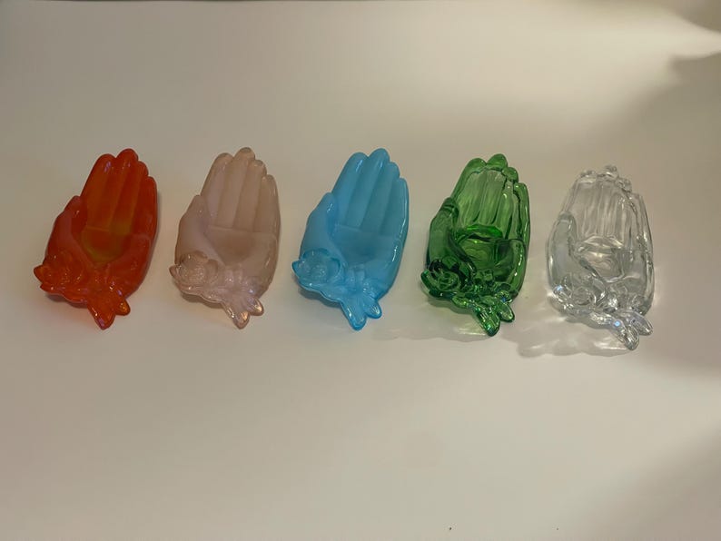 May include: Five glass hand-shaped trinket dishes in various colors. The dishes are arranged in a row from left to right: orange, pink, blue, green, and clear.