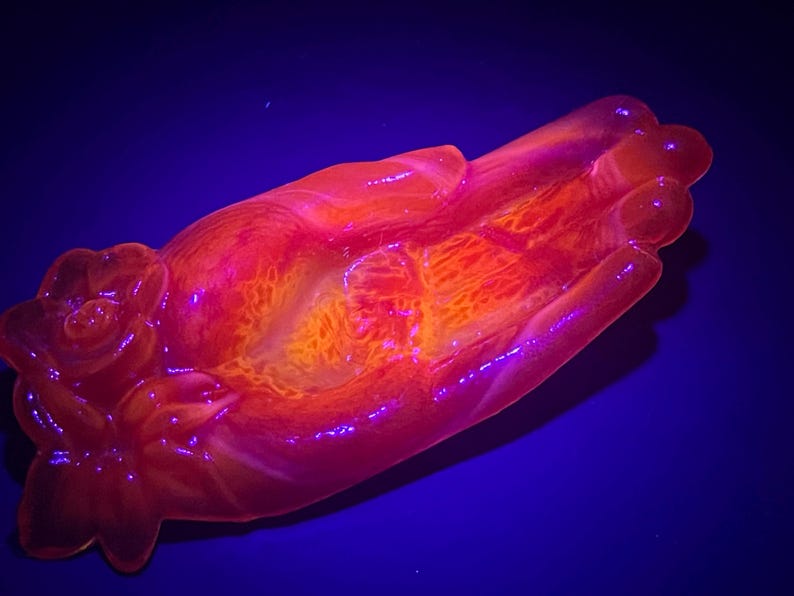 May include: A hand-shaped dish made of resin with a rose design on one end. The dish is a vibrant orange and pink color and glows under a black light.