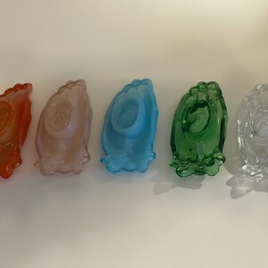 May include: Five glass dishes in different colors, including orange, pink, blue, green, and clear. Each dish has a raised design that resembles a pair of hands clasped together in prayer.