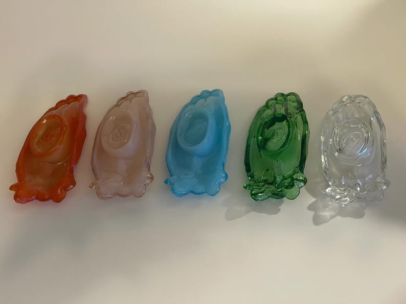 May include: Five glass dishes in different colors, including orange, pink, blue, green, and clear. Each dish has a raised design that resembles a pair of hands clasped together in prayer.