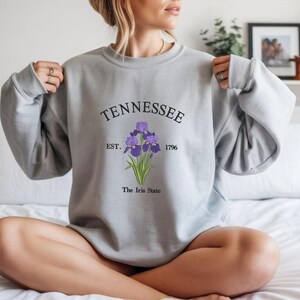 May include: A light gray sweatshirt featuring the word "Tennessee" above a purple iris flower illustration. Below the flower, the text reads "The Iris State" and "Est. 1796". The sweatshirt has long sleeves and a crew neck.