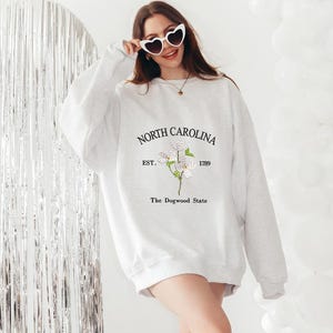 Pu&ograve; includere: Una felpa grigio chiaro con la scritta "NORTH CAROLINA EST. 1789" e una grafica floreale. La felpa ha anche il testo "The Dogwood State". La persona indossa occhiali da sole a forma di cuore e stivali da cowboy.